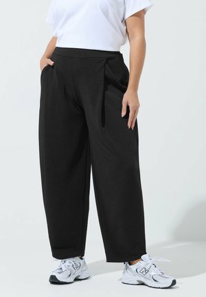 Person trägt schwarze Hosen mit weitem Bein, weißes Kurzarmoberteil und weiße New Balance Sneaker und steht mit den Händen in den Taschen.