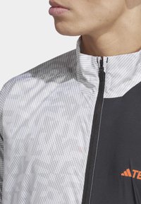 Leichte Jacke mit einem grauen, vertikal gestreiften Muster auf einer Seite und einfarbig schwarz auf der anderen. Verfügt über einen hohen Kragen und einen Reißverschluss.