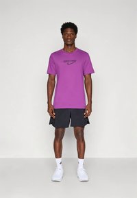 Camiseta morada Nike Pro con logo negro, combinada con pantalones cortos deportivos negros. El modelo lleva zapatillas blancas y calcetines, y está de pie sobre un fondo liso.