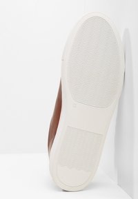 Braune Leder-Sneaker mit einer weißen Gummisohle. Die Sohle ist mit einem Wellenmuster für besseren Halt ausgestattet, und der Schuh hat eine glatte Oberfläche.