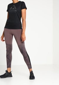 Černý běžecký tričko Reebok, spárované s hnědými a černými legínami s vzorem, s elegantním designem a strukturovanou tkaninou. Černé sportovní boty.