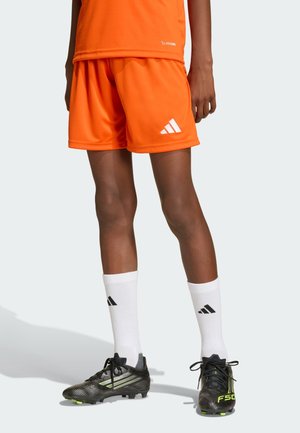 Person wearing bright orange athletic shorts and top, white Adidas crew socks, and black soccer cleats with yellow accents.