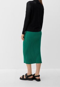 Schwarzer Strickpullover, grüner gerippter Midirock und schwarze Sandalen mit breiten Riemen. Fokus auf Texturvariation und Farbkontrast.