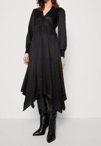 Robe noire à manches longues, plissée, avec un décolleté en V et un ourlet asymétrique. Portée avec des bottes noires montantes, à bout pointu.