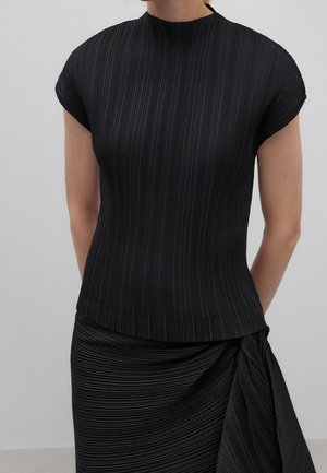 Kobieta ubrana w czarny, prążkowany top z krótkim rękawem i dopasowaną plisowaną spódnicę z marszczeniem po prawej stronie.