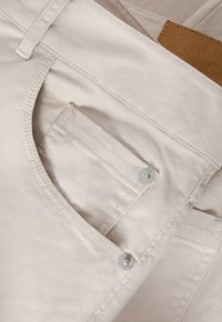 Beige nadrág, elülső zsebével, amely patentos záródással rendelkezik, és sima, twill szerkezettel készült. Az öltések világosak és egyformák.