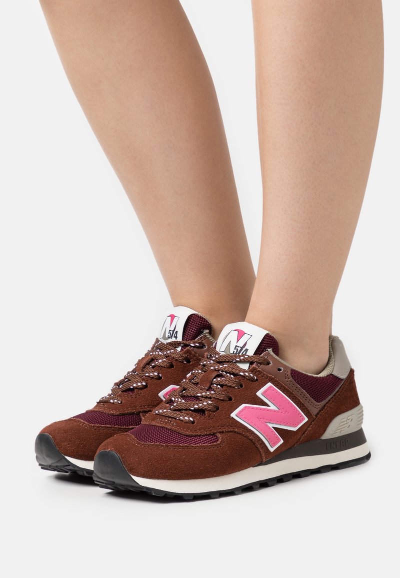 Baskets marron et bordeaux avec un logo rose "N" portées pieds nus sur fond blanc.