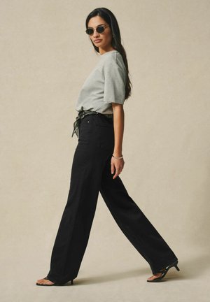 Naine, kes kannab ümarate klaasidega päikeseprille, halli T-särki, musti laia säärega pükse ja musti kontsaga sandaale, kõnnib külgsuunas beeži tausta ees.