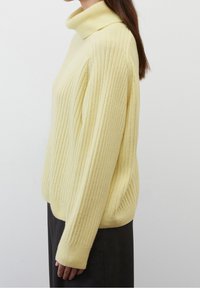 Pullover giallo a coste con colletto alto e maniche lunghe, caratterizzato da una vestibilità rilassata e da un morbido tessuto, indossato sopra una gonna scura.