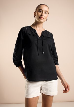 Femme portant un blouse noire avec des détails en dentelle et un short blanc retroussé, se tenant devant un fond beige uni.