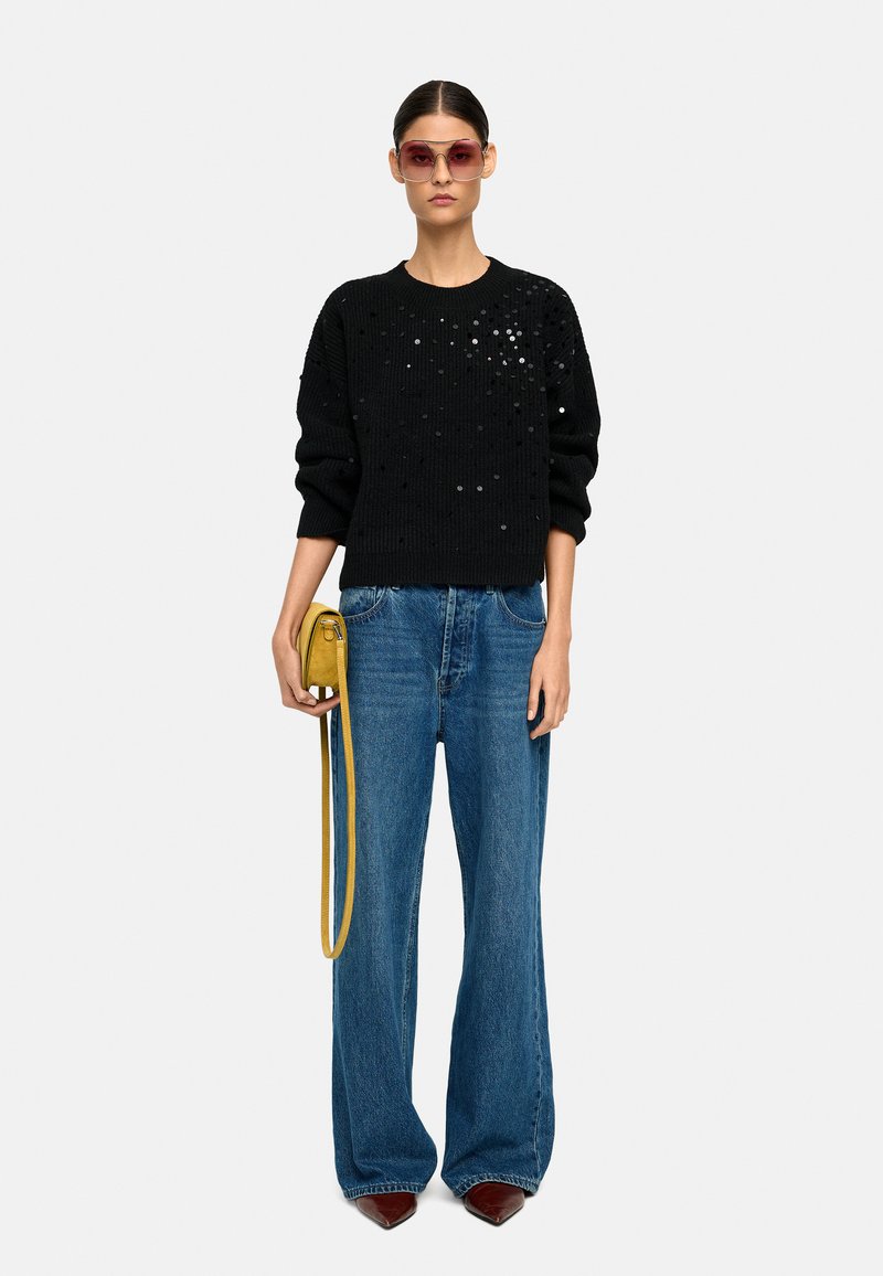 Czarny sweter na drutach z cekinami, niebieskie szerokie dżinsy, mała żółta torebka na ramię i fioletowe okulary przeciwsłoneczne. Gładkie tekstury i odważne kolory.