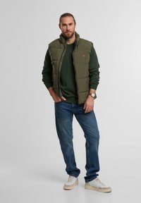 Mężczyzna z brodą i związanymi włosami, ubrany w zielony sweter, oliwkową kurtkę puchową, niebieskie dżinsy, białe sneakersy i zegarek na nadgarstku, stojący na jednolitym tle.