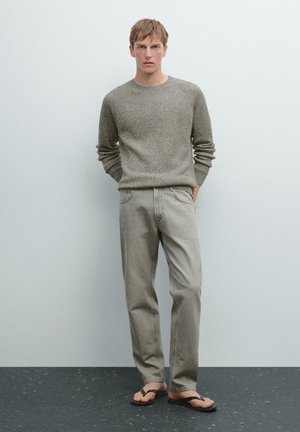 Light brown, textured knit sweater, gray straight-leg jeans, and brown flip-flops. Model stands with hands in pockets against a neutral background.