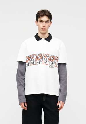 Ung mand iført en hvid polo med sort krave og orange blomsterprint, lagdelt over en grå langærmet skjorte og sorte bukser.