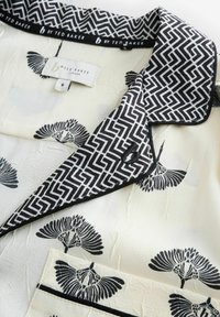 Schwarz-weiße gemusterte Bluse aus strukturiertem kremfarbenem Stoff, mit einem geometrischen Kragen und Vogelmotiven, Etikett unter dem Kragen angebracht.