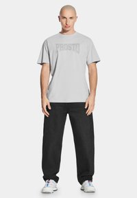 Jasnoszara bawełniana koszulka z nadrukiem "PROSTO" w ciemniejszym odcieniu, zestawiona z czarnymi workowatymi dżinsami i białymi sneakersami.