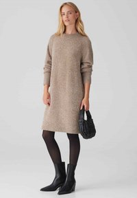 Abito maglione in beige con una finitura testurizzata, maniche lunghe e una vestibilità rilassata, abbinato a stivali neri alla caviglia e una borsa testurizzata.