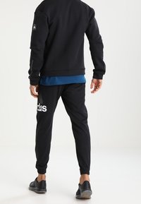 Sweatshirt noir avec un t-shirt bleu en dessous, pantalon de survêtement noir avec le logo "adidas" blanc, et chaussures de sport noires.