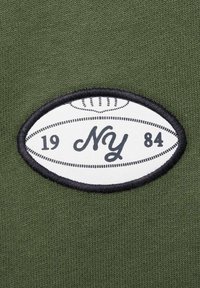 Tessuto verde con finitura texturizzata, dotato di una toppa ovale bianca. La toppa ha un bordo nero e il testo "NY 1984" in scrittura nera. Include un grafico di un pallone da football.