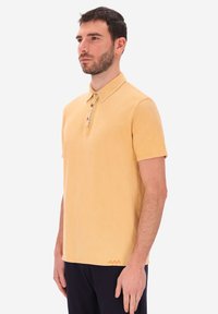 Uomo con barba che indossa una polo gialla a maniche corte e pantaloni scuri, in piedi di profilo contro uno sfondo bianco semplice.