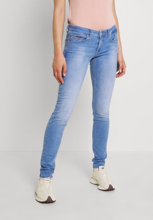 Mujer vestida con jeans ajustados de color azul claro y zapatillas blancas, de pie frente a un fondo liso claro.