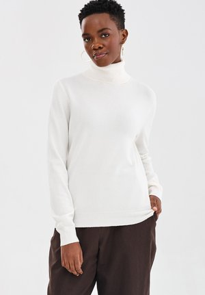 Person trägt einen weißen Rollkragenpullover und eine dunkelbraune Hose, steht mit einer Hand in der Tasche vor einem schlichten Hintergrund.