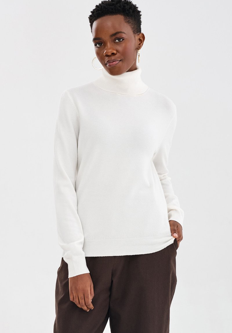 Osoba w białym swetrze z wysokim kołnierzem i ciemnobrązowych spodniach, stojąca z jedną ręką w kieszeni na jednolitym tle.