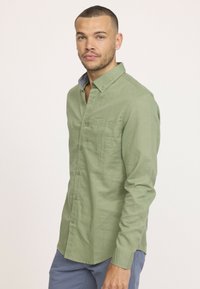 Chemise à boutons en lin vert clair avec manches longues, une poche poitrine et une doublure de col bleue contrastante. Texture lisse, design décontracté.