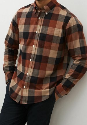 Mężczyzna ubrany w koszulę z długim rękawem zapinaną na guziki, z dużym kratkowanym wzorem w brązowych, beżowych, czarnych i rdzawych kolorach, z jedną ręką w kieszeni.