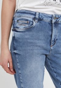 Primer plano de una persona que lleva jeans de mezclilla azul claros y una camisa blanca, con la mano relajada a su lado.