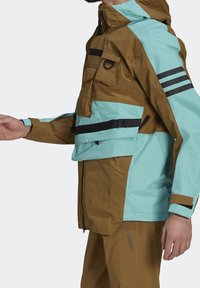 Personne portant une veste d'extérieur marron et bleu clair avec plusieurs poches, fermetures éclair et des bandes noires sur la manche, ainsi qu'un pantalon marron.