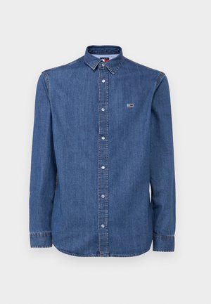 Camisa de mezclilla abotonada en azul oscuro con cuello, mangas largas y un pequeño logo en el pecho. Presenta costuras de contraste y cierre de botones.