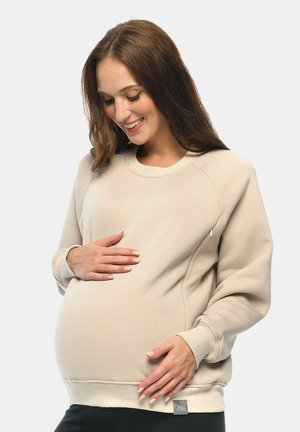 Schwangere Frau lächelt, trägt einen beigen Pullover und hält sanft mit beiden Händen ihren Babybauch vor einem hellen Hintergrund.
