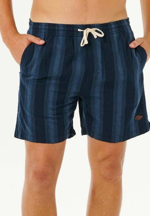 Hombre vistiendo pantalones cortos de baño azul marino con un patrón de rayas verticales y cordón blanco, con las manos en los bolsillos.