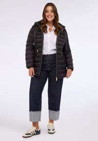 Chaqueta acolchada negra con capucha, con cierre frontal y detalles en dorado. Combinada con vaqueros azul oscuro y zapatillas crema.