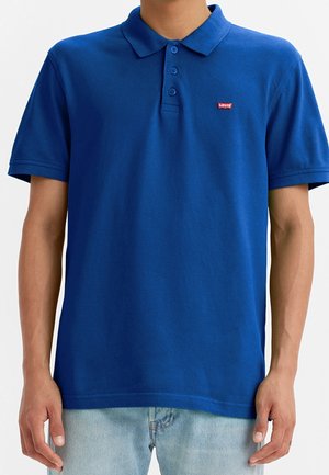Mężczyzna ubrany w niebieską koszulkę polo Levi's z trzema guzikami i jasnoniebieskie dżinsy, stojący na jednolitym tle.