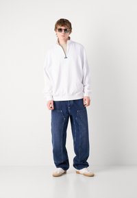 Pull blanc avec demi-zip et poignets côtelés, associé à un jeans bleu marine ample avec poches avant et des baskets blanches.