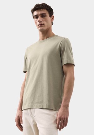 Giovane uomo che indossa una t-shirt a maniche corte di colore verde oliva chiaro tinta unita e pantaloni color avorio, in piedi davanti a uno sfondo bianco.