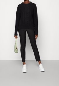 Mustavillattu neule, jossa on tekstuuripintoja, yhdistetty hoikkanahkiapuntoihin ja valkoisiin tennareihin. Kantaa vaaleanvihreää käsilaukkua, jossa on hihna.