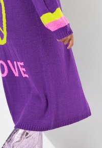 Violetti neulemekko, jossa on ylipitkä malli; siinä on "LOVE" vaaleanpunaisena ja graafinen yksityiskohta neonkeltaisena. Ribbattu helma ja hihansuut.