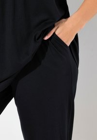 Haut en tissu noir et doux avec une poche ; vue rapprochée montrant une texture lisse et une coupe décontractée, avec une main placée dans la zone de la poche.
