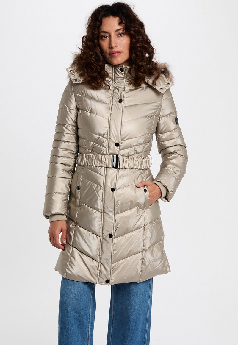 Beige pufferjas met een ceintuur in de taille, een chevron patroon en een afneembare capuchon met bontafwerking. Voorzien van drukknoopsluitingen en zijzakken.