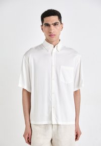 Chemise blanche à manches courtes en tissu lisse avec une poche sur le côté gauche et un col classique, portée avec un pantalon beige clair.
