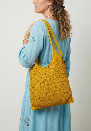 Vrouw met golvend haar, gekleed in een blauwe geborduurde jurk, draagt een grote gele handtas met bloemvormige uitsnijdingen over haar schouder.