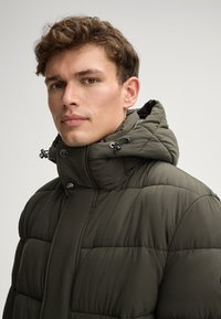 Giacca imbottita di colore verde scuro, con colletto alto e cappuccio staccabile, caratterizzata da accessori neri, cuciture visibili e una texture liscia.