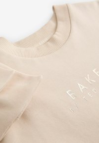 Měkká béžová mikina s žebrovaným výstřihem. Na přední straně se nachází kovový nápis "BAKERS BY TED" a hladká strukturovaná látka.