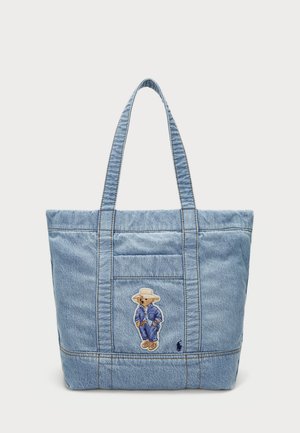Bolso de lona de mezclilla con dos correas largas, que presenta un oso bordado con un traje azul y un sombrero de paja en la parte frontal, con detalles de costura visibles.