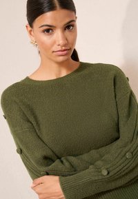 Pulover verde tricotat cu gât rotund, material texturat și accente circulare de-a lungul mânecilor. Material moale și călduros.