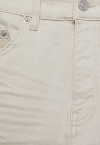 Primer plano de unos jeans de denim color crema que muestran el bolsillo, el lazo del cinturón, los detalles de costura y el botón y remaches de metal plateado.