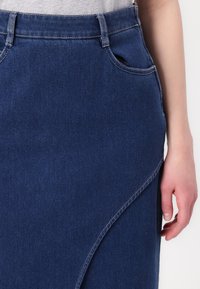 Τζιν φούστα σε σκούρο μπλε χρώμα, με εφαρμοστό μέσο, πλαϊνές τσέπες και ασύμμετρο στρίφωμα με αντίθετα ραφές.